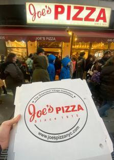 A Joe's Pizza box held in front of the restaurant with a line of people waiting.