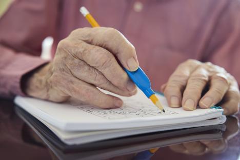 Close-up of a senior man's arthritic hands using a grip aid to write in a Sudoku puzzle book.