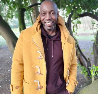 A smiling man in a mustard-yellow coat stands near a tree.