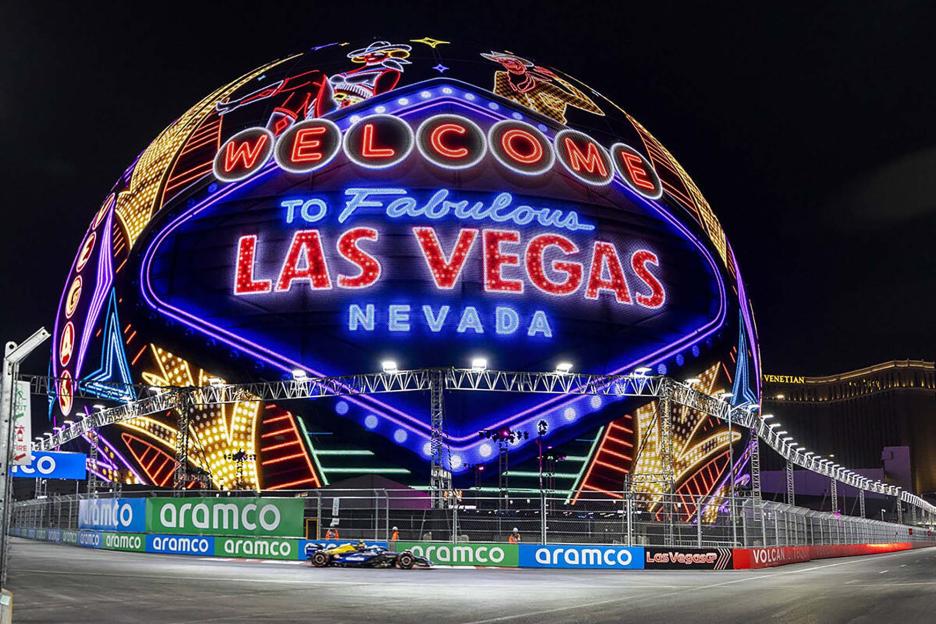 A Formula 1 race car on the track with the Sphere displaying "Welcome to Fabulous Las Vegas Nevada" in neon lights in the background.