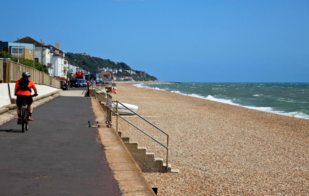 Person cycling along a paved path next to a pebble beach and ocean.