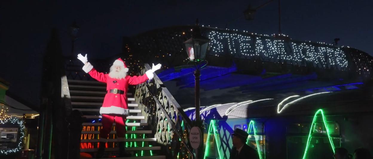 Santa Claus standing on steps decorated with Christmas lights, with a train car illuminated with "STEAMLIGHTS" in the background.