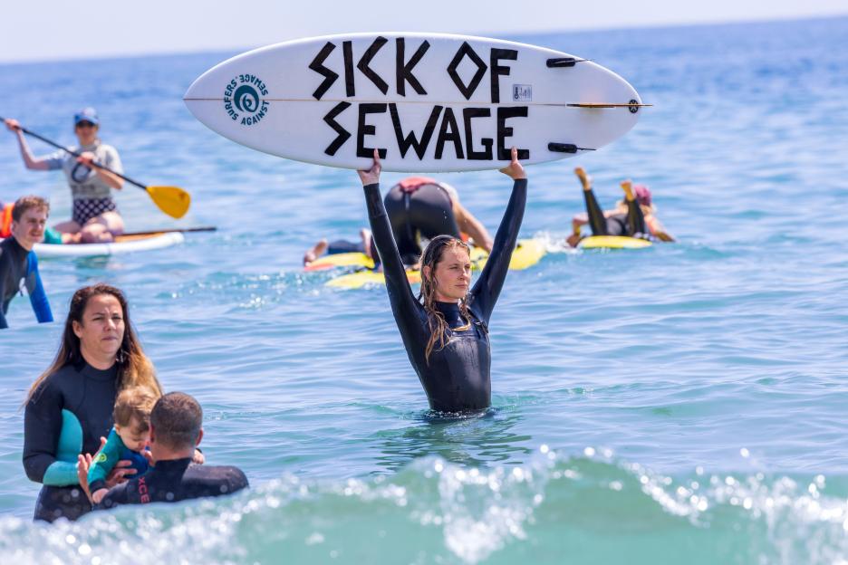 Protestor holding a surfboard with "Sick of Sewage" written on it during a beach protest.