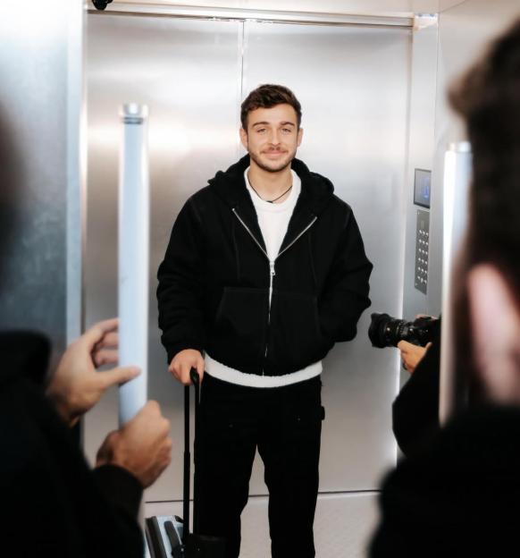 George Clarke in an elevator with luggage, being photographed.
