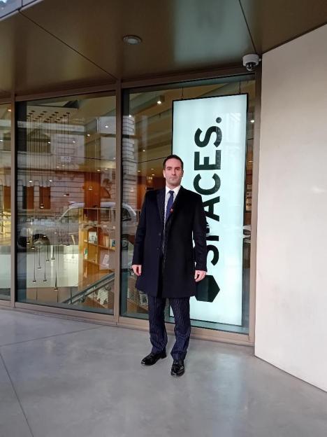 A man in a dark coat and pinstriped trousers stands outside a "SPACES." building.