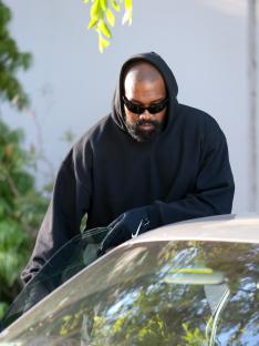 Kanye West wearing a black hoodie and sunglasses, leaning on a car.