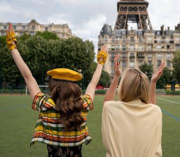 Two women with their backs to the camera, wearing yellow gloves and celebrating in front of the Eiffel Tower.