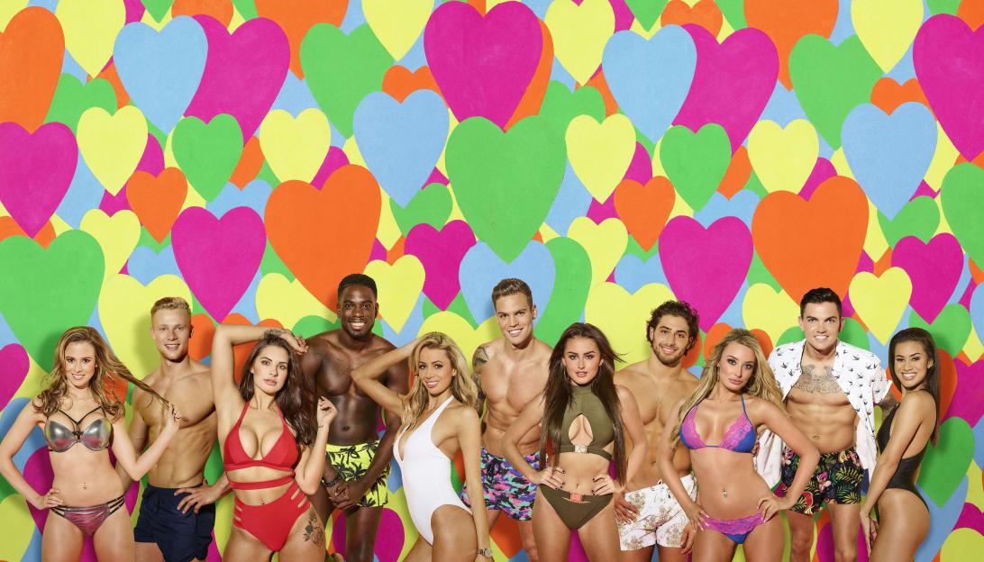 Love Island contestants Harley, Marcel, Dominic, Kem, Sam, Olivia, Jessica, Camilla, Amber, Chloe, and Montana pose in front of a colorful heart background.