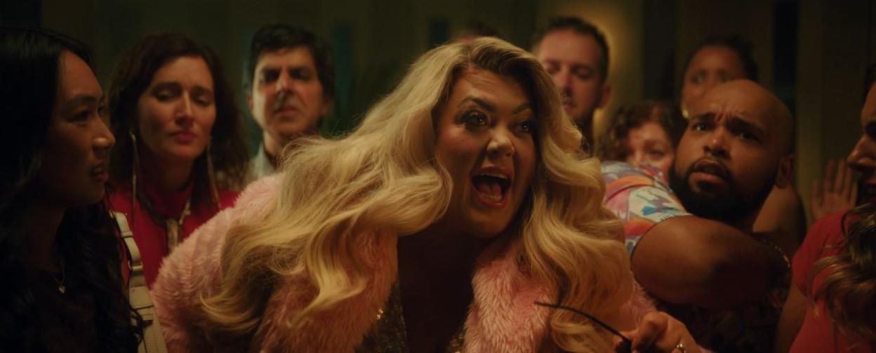A person with long blonde hair and a pink fur coat, mouth open, looking shocked among a crowd of people.
