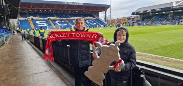 Two boys at a football stadium holding an Ackley Town FC scarf and a model FA Cup trophy.