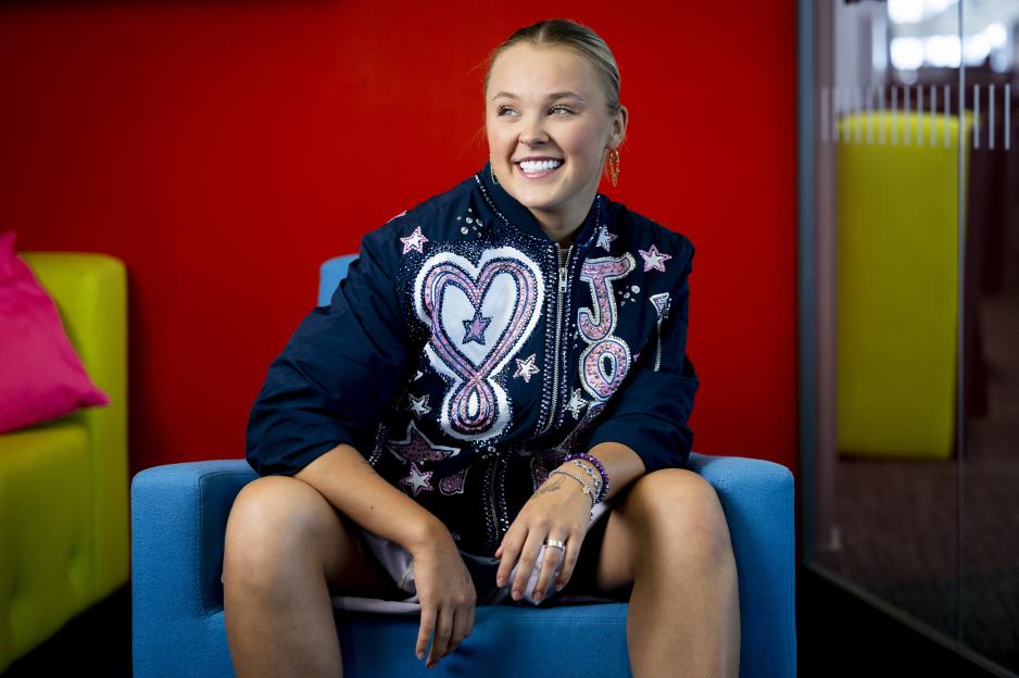 JoJo Siwa sitting in a blue chair, smiling and looking to the right.