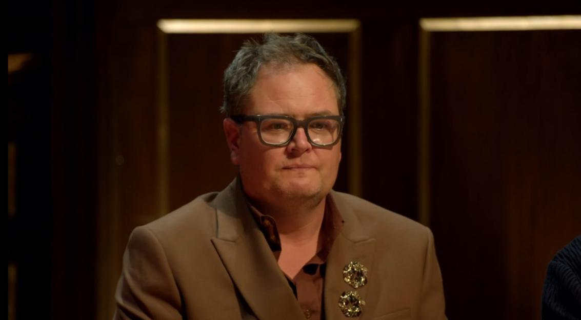 A man in a brown suit with two brooches on the lapel, wearing black-rimmed glasses.