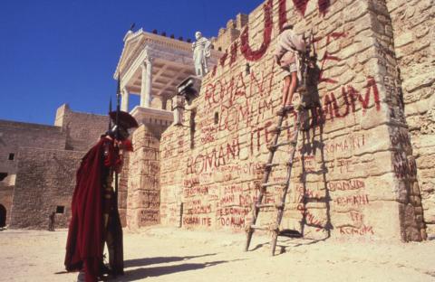 A still from Monty Python's Life of Brian showing a wall with graffiti and a Roman soldier.