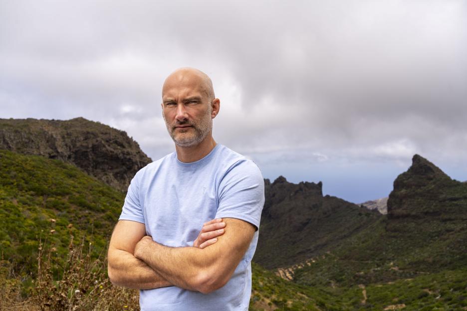 A man with arms crossed stands in front of a mountainous landscape.