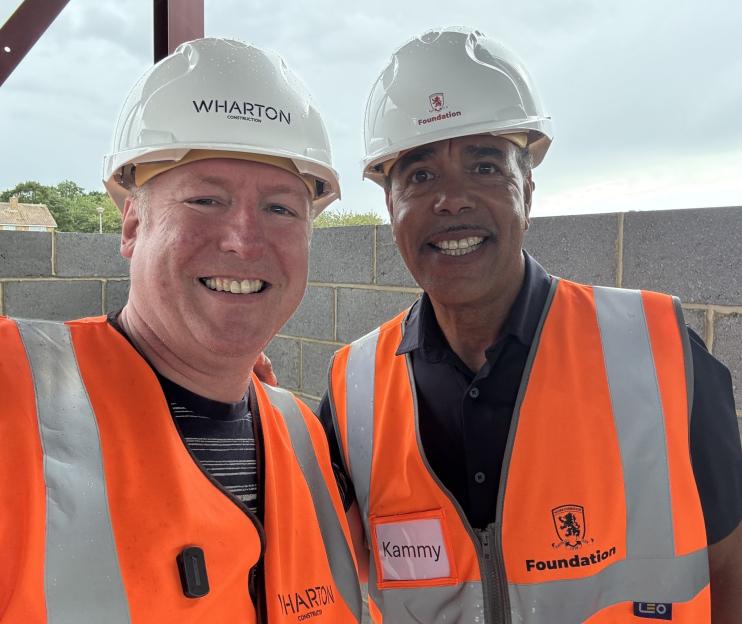 Two men in hard hats and safety vests smile for a photo at a construction site.