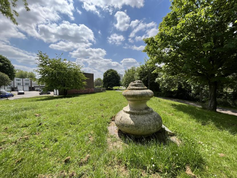 An outdoor concrete bollard on a grassy hill with trees and buildings in the background.