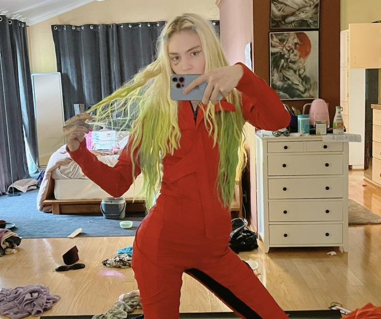 Grimes, in a red jumpsuit with black stripes, takes a mirror selfie while holding up her blonde and green hair.
