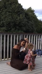 A family on a deck; a pregnant woman and a man are seated with their toddler daughter, who is holding a bouquet of flowers.