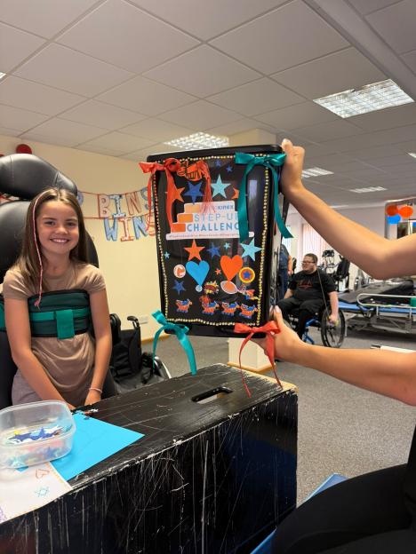 Ivy Amos, 9, smiling and wearing a harness, stands next to a decorated black box held by two people.