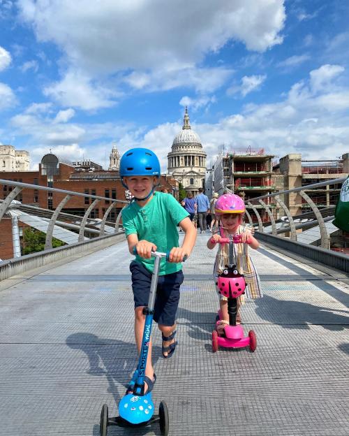 Two children riding scooters on a bridge with St. Paul's Cathedral in the background.
