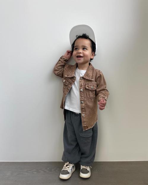 Tatum Thompson standing against a white wall wearing a white shirt, brown jacket, gray sweatpants, and white sneakers, with a white hat on his head.