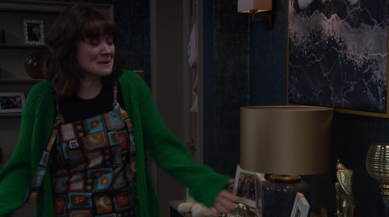 Screenshot of a woman wearing a green cardigan and patterned overalls, appearing upset.