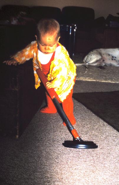 A toddler holding a vacuum cleaner handle while standing on a rug.