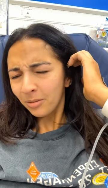 A woman with dark hair grimacing in discomfort, with hospital equipment visible in the background.