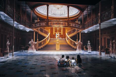 A family sits on the floor facing a digital recreation of the Titanic's Grand Staircase, with translucent figures of passengers dancing and mingling.