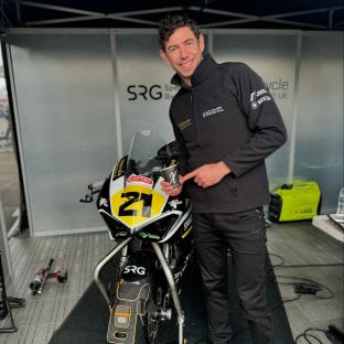 Man in racing jacket holding coffee cup next to motorcycle.