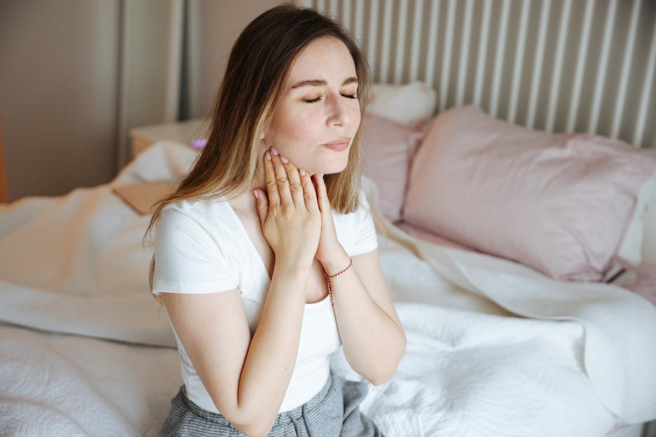 Woman sitting on bed holding her throat.