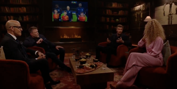Screenshot of four people sitting in armchairs around a coffee table, watching a soccer game on a television screen.