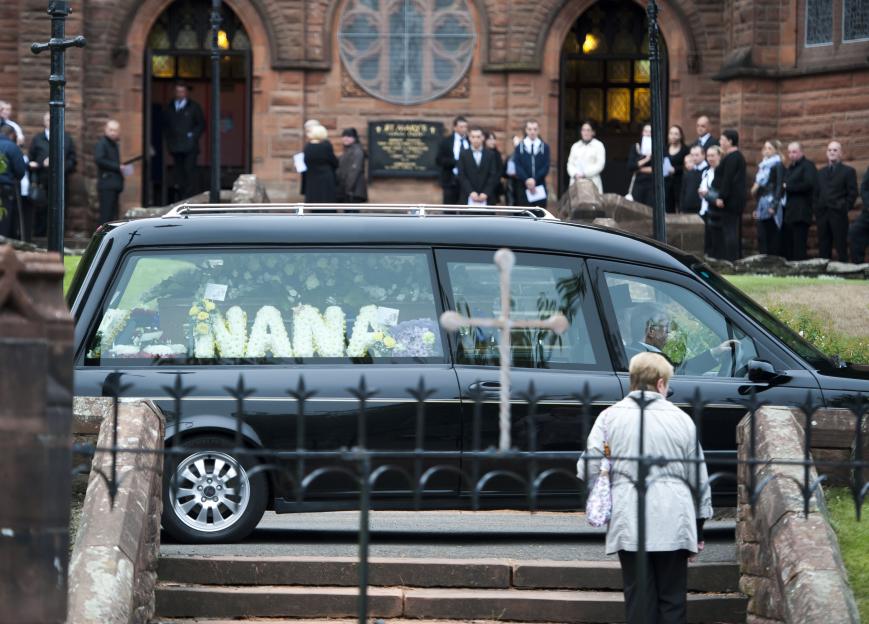 Hearse with floral arrangement spelling "Nana" arriving at a church.