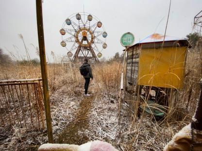 Person standing in snow-covered overgrown area of abandoned theme park with Ferris wheel in background.