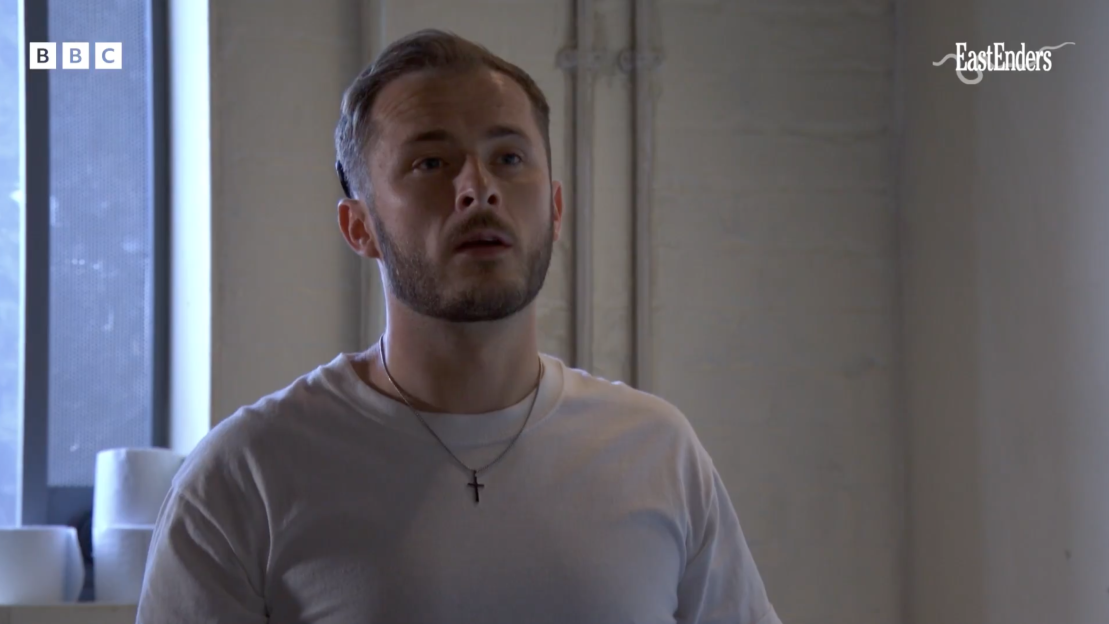 A young man with a beard and light brown hair looks up in a still from the show EastEnders.