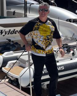 Man in patterned shirt standing by a yacht and inflatable boat.