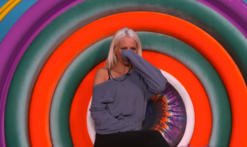 A woman with blonde hair in an off-the-shoulder top covers her mouth and nose with her sleeve while crying.