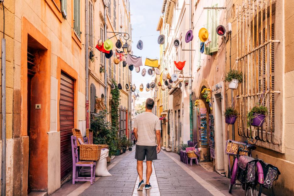 A man walks down a narrow, colorful street with hats and clothes hanging overhead.