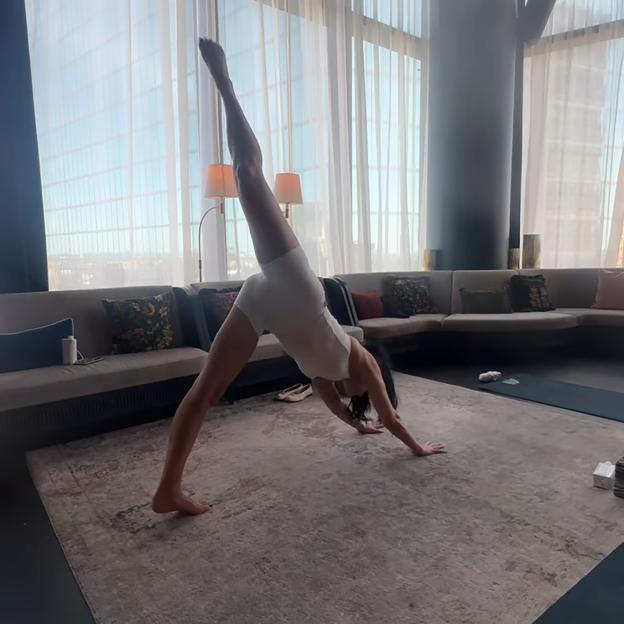 Dua Lipa in a white leotard performing a yoga pose, lifting one leg straight into the air in a downward dog variation.