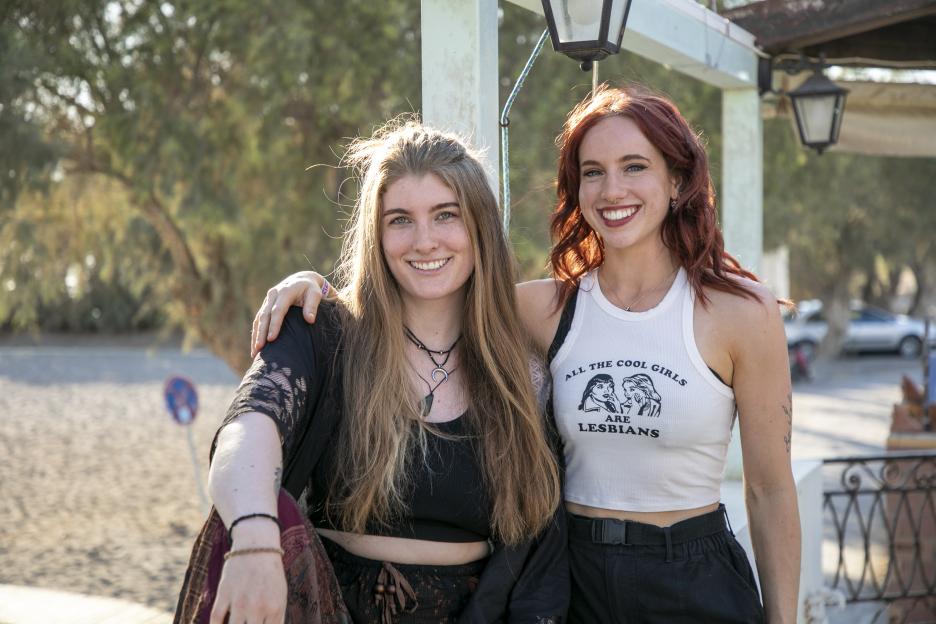 Two smiling girlfriends, one with long blond hair and the other with red hair wearing a tank top that reads "All the cool girls are lesbians."