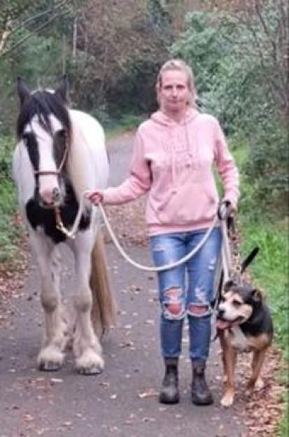 Leanne Williams, a woman in a pink hoodie and ripped jeans, stands between a black and white horse and a brown and black dog on a path.