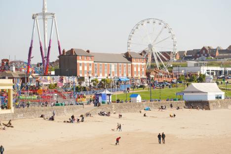 People enjoying a sunny day at a beach with amusement park rides and a Ferris wheel in the background.