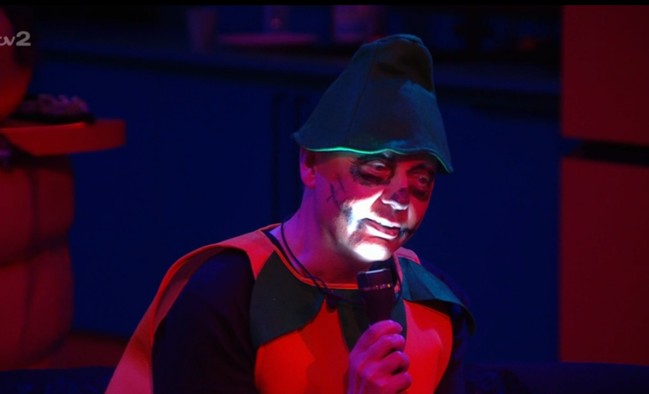 A person wearing an orange and green costume, face painted, holding a microphone.