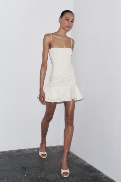 Model wearing a white ruffled mini dress and white heels.