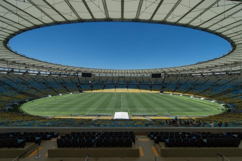 An empty football stadium with blue and yellow seats under a clear blue sky, viewed from a shaded seating area.
