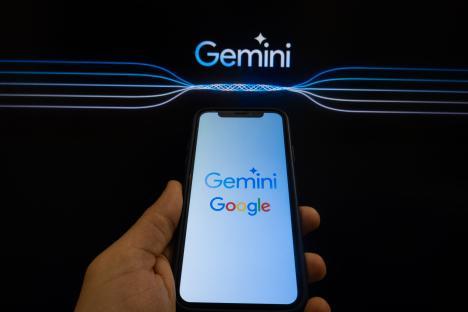 Photo illustration of a hand holding a smartphone displaying the Gemini Google logo, with the Gemini logo also displayed on a screen in the background.