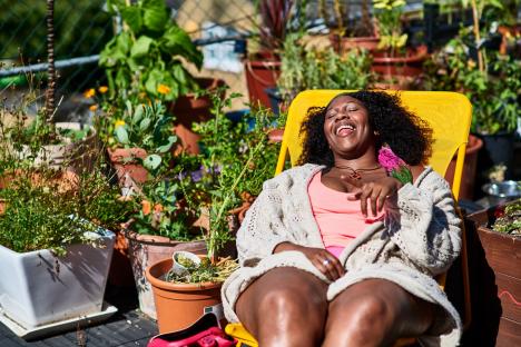 Black woman in her 30s sitting on patio chair on rooftop garden, enjoying the sun, carefree, leisure, weekend