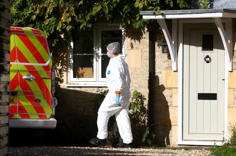 A forensic officer in a white protective suit and face mask walks past a police van with orange and yellow stripes outside a stone building with a light-colored door.