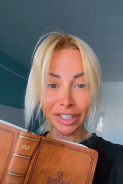 A blonde woman holding a brown book with a cross on the cover.