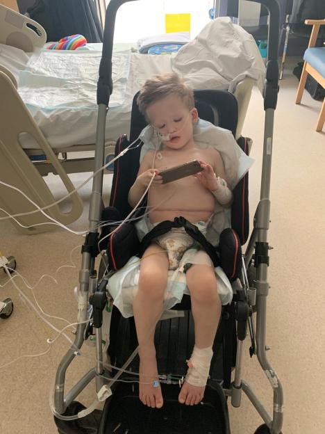 A young boy, Tommy Verbickas, in a hospital chair, has medical tubes and wires attached to him while looking at a phone.